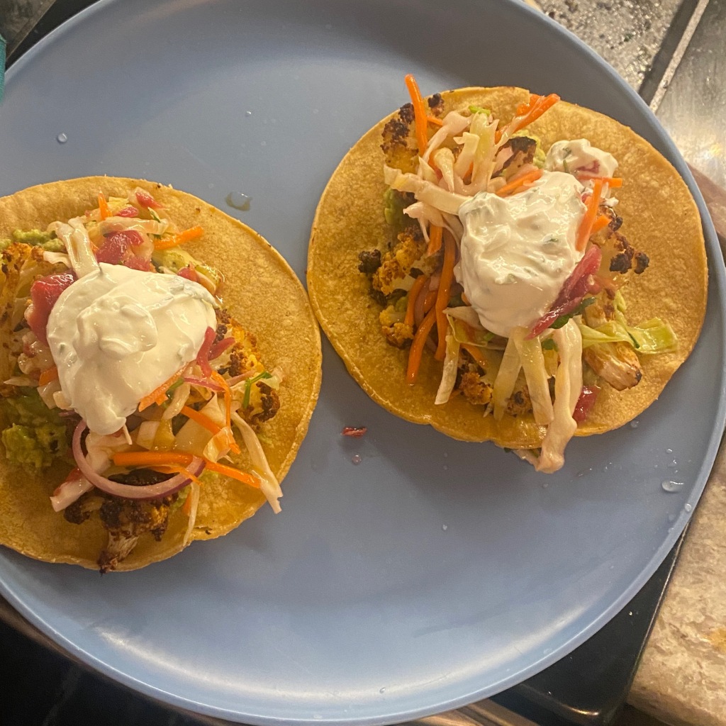two tacos sit on a blue plate. They are composed of corn tortillas with guacamole, chipotle roasted cauliflower, a cabbage slaw, and topped with a cilantro lime crema.